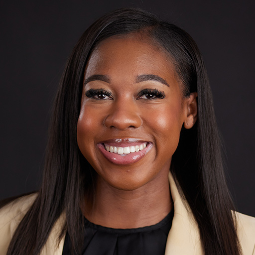 A woman smiling brightly, wearing a black blazer and white top with elegant drop earrings, is introduced against a dark background—Meet The Team At GGRM.