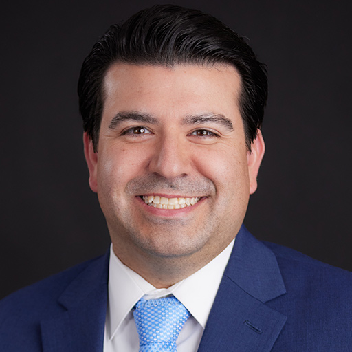 Meet The Team At GGRM: A man with dark hair wearing a navy blue suit, white shirt, and light blue tie, smiling in front of a dark gray background.