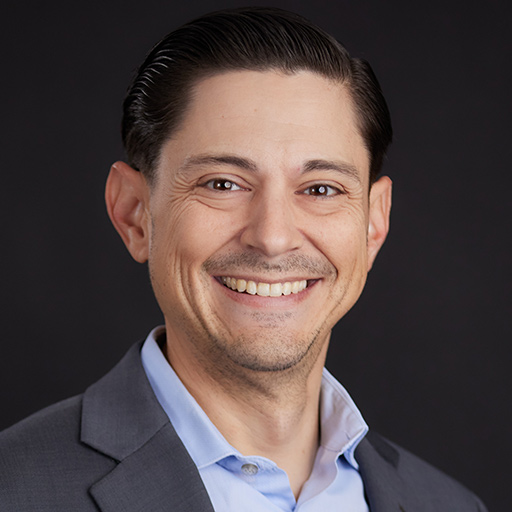Meet The Team At GGRM: A man with dark hair wearing a blue suit, light blue shirt, and a colorful patterned tie smiles at the camera against a plain dark background.