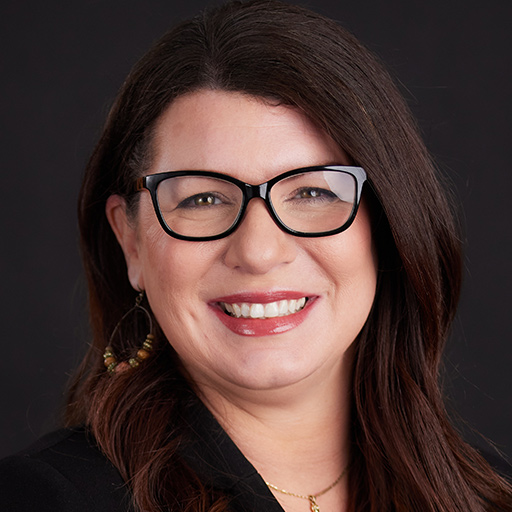 A woman with long brown hair and glasses smiles at the camera. She is wearing a black blazer and posing in front of a dark gray background as part of the Meet The Team At GGRM series.