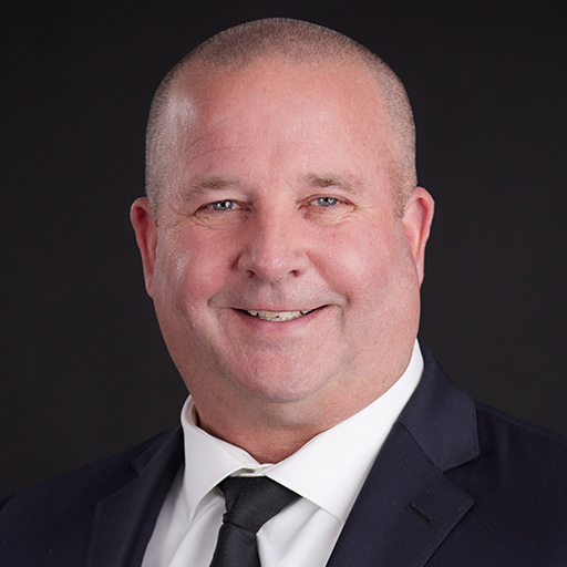 Meet The Team At GGRM: A middle-aged man with short hair, wearing a gray suit, white shirt, and blue polka dot tie, smiles at the camera against a dark gray background.