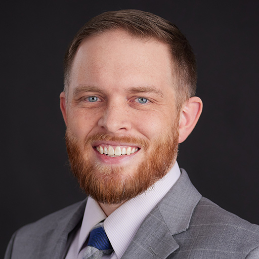 Meet The Team At GGRM: A man with light skin, short brown hair, and a beard is smiling. He is wearing a gray suit, white shirt, and a purple polka dot tie, with a dark background.