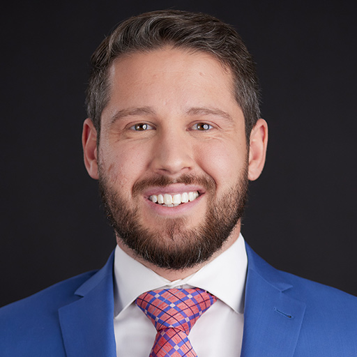 Meet The Team At GGRM: A man with short brown hair and a beard, wearing a blue suit, white shirt, and red patterned tie, smiles in front of a plain dark background.