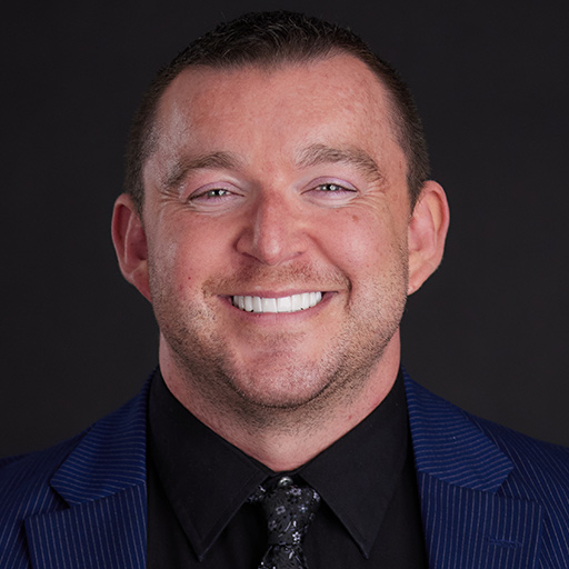 Smiling man with short brown hair wearing a dark suit, white dress shirt, and patterned tie, posed in front of a plain dark background. Meet The Team At GGRM.