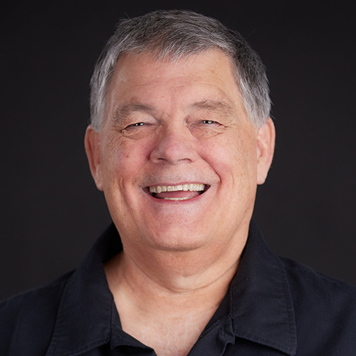 A middle-aged man with short gray hair smiles at the camera. He is wearing a gray shirt over a black top, set against a dark, plain background. Meet The Team At GGRM and discover the friendly faces behind our expertise.