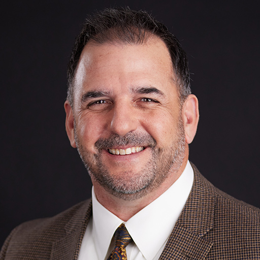 Meet The Team At GGRM: Smiling man in a light blue plaid suit, white shirt, and patterned tie stands in front of a dark gray background.