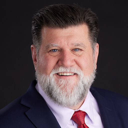 Meet The Team At GGRM: A middle-aged man with a gray beard and brown hair wears a dark suit, white shirt, and purple tie, smiling warmly in front of a plain dark background.