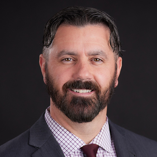 Meet The Team At GGRM: A man with short dark hair, a trimmed beard, and mustache, wearing a gray suit, white shirt, and red tie, smiles in front of a dark gray background.