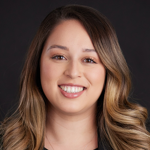 Meet The Team At GGRM: A woman with long brown hair, wearing a dark blazer over a white blouse, smiles at the camera against a plain dark background.