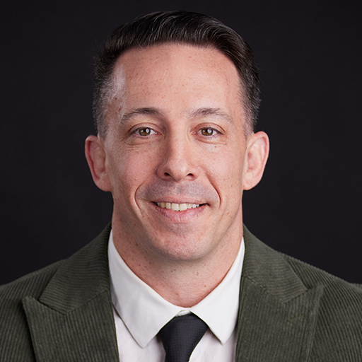 Meet The Team At GGRM: A man wearing a rust-colored suit jacket, white dress shirt, and matching tie, posing for a professional headshot against a dark gray background.