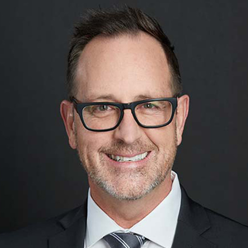 Smiling man with short brown hair, wearing dark-rimmed glasses, a dark suit, white shirt, and striped tie, posed against a plain dark background. Meet The Team At GGRM.