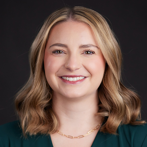 Meet The Team At GGRM: A woman with straight, light brown hair wearing a dark green top, large sunburst earrings, and a matching sunburst necklace smiles in front of a dark, plain background.