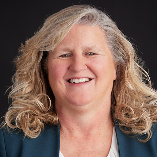 Meet The Team At GGRM: A middle-aged woman with wavy blonde hair, wearing a teal blazer and white blouse, smiles warmly in front of a plain dark background.