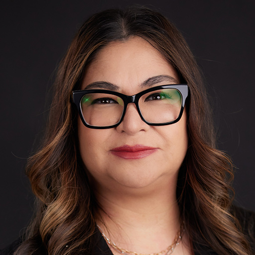 Smiling woman with long wavy brown hair, wearing black glasses, gold hoop earrings, layered gold necklaces, and a light green jacket, posing in front of a plain dark background. Meet The Team At GGRM.