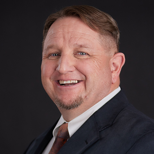 Smiling man with short light brown hair, wearing a navy suit, blue striped dress shirt, and a patterned yellow-and-blue tie, posed in front of a dark gray background. Meet The Team At GGRM.