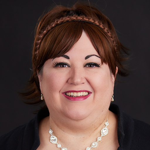 Meet The Team At GGRM: A woman with dark hair pulled back, wearing a gold headband and black top, smiles broadly against a plain dark background.