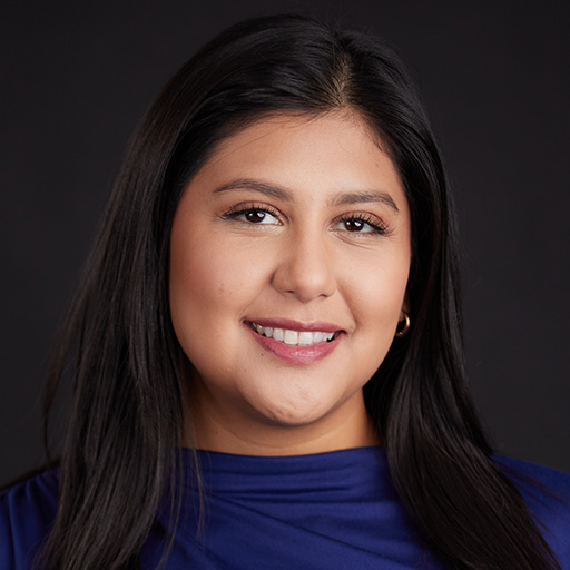 Meet The Team At GGRM: A woman with long, straight, dark hair and medium skin tone is smiling in a blue top, posed in front of a dark background.