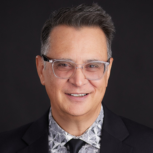 Meet The Team At GGRM: A man wearing glasses, a gray suit, white shirt, and patterned tie smiles in front of a dark gray background.