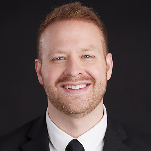 Meet the Team at GGRM: A man with short light brown hair and a trimmed beard smiles at the camera, wearing a black shirt against a plain dark background.