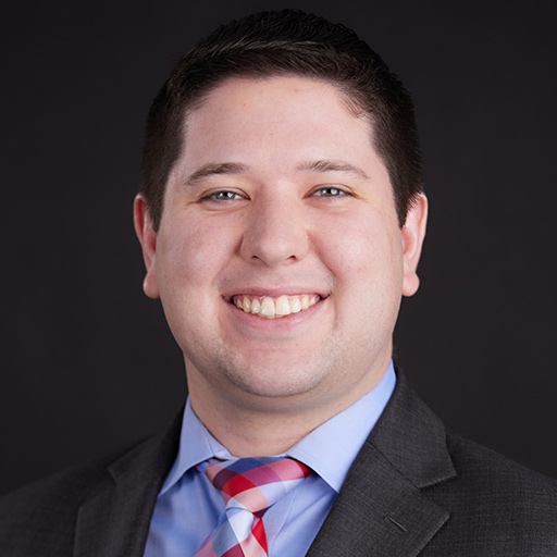 Meet The Team At GGRM: A young man wearing a dark suit, light blue shirt, and plaid tie smiles at the camera. He has short brown hair, an American flag pin on his lapel, and stands against a plain, dark background.