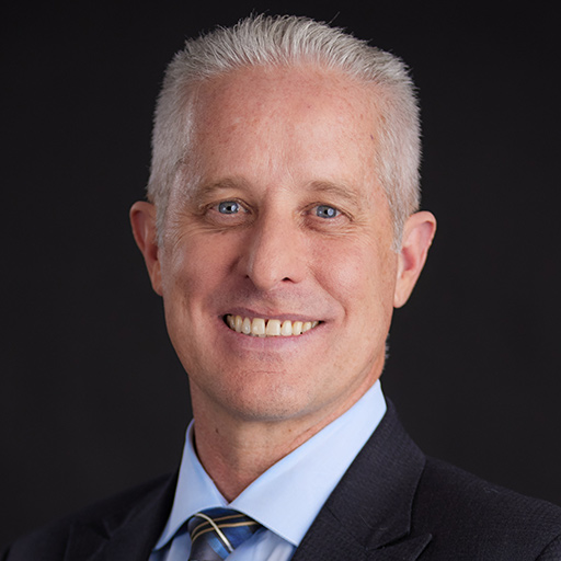 Smiling man with short gray hair wearing a dark suit, light blue dress shirt, and blue patterned tie, posing in front of a dark gray background. Meet The Team At GGRM.