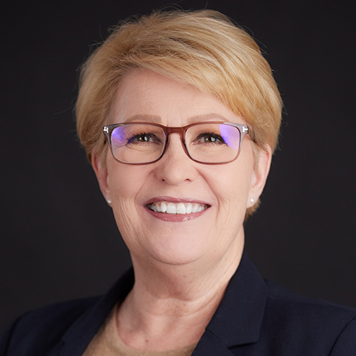 Meet the Team at GGRM: A smiling older woman with short, light blonde hair, wearing clear glasses, a beige blazer, and a light-colored scarf, poses against a dark gray background.