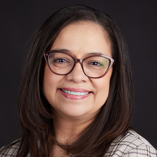 Meet The Team At GGRM: A woman with curly dark hair and glasses smiles softly, wearing a black and white patterned top and a delicate necklace, posed against a plain dark background.
