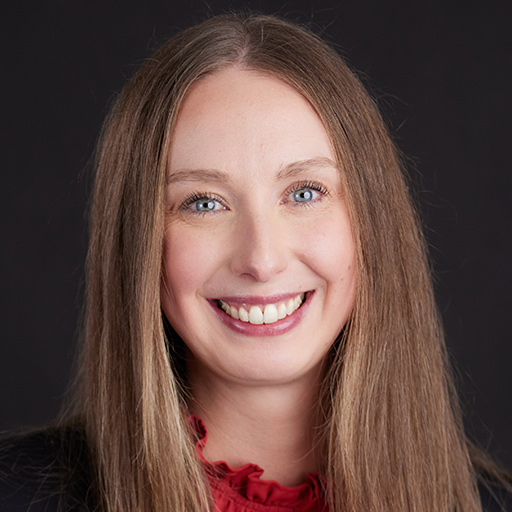 Meet the team at GGRM: A woman with long straight light brown hair smiles at the camera. She is wearing a black top and a red blouse underneath, photographed against a plain dark background.