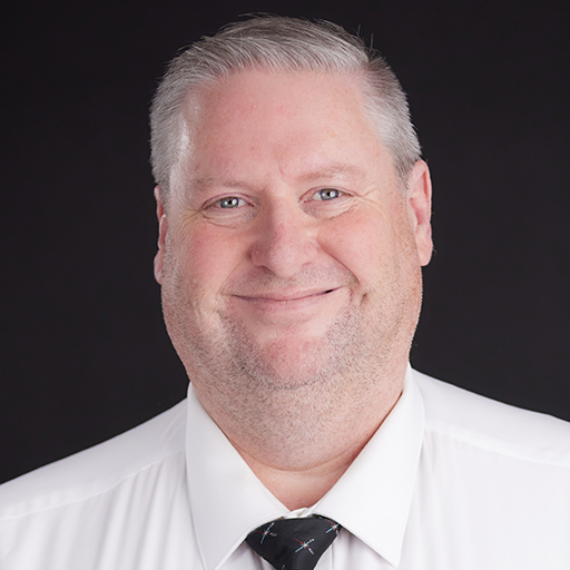 Meet the Team at GGRM: A middle-aged man with short gray hair smiles while wearing a white dress shirt and a black patterned tie, posed against a plain dark background.