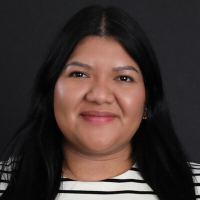 Meet The Team At GGRM: A woman with long dark hair and brown skin smiles at the camera. She is wearing a black and white striped top and is posed against a plain dark background.