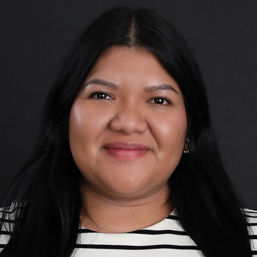 Meet The Team At GGRM: A woman with long dark hair and brown skin smiles at the camera. She is wearing a black and white striped top and is posed against a plain dark background.