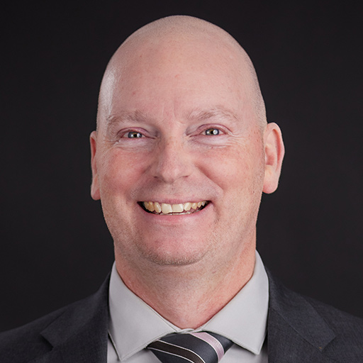 Meet The Team At GGRM: A bald man in a dark suit, light shirt, and striped tie smiles at the camera against a plain dark background.